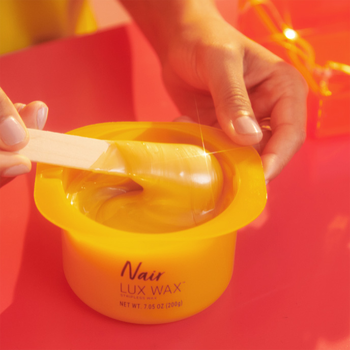 A woman's hands dipping a spatula into a Nair Lux Wax™ pot filled with warm, creamy wax.